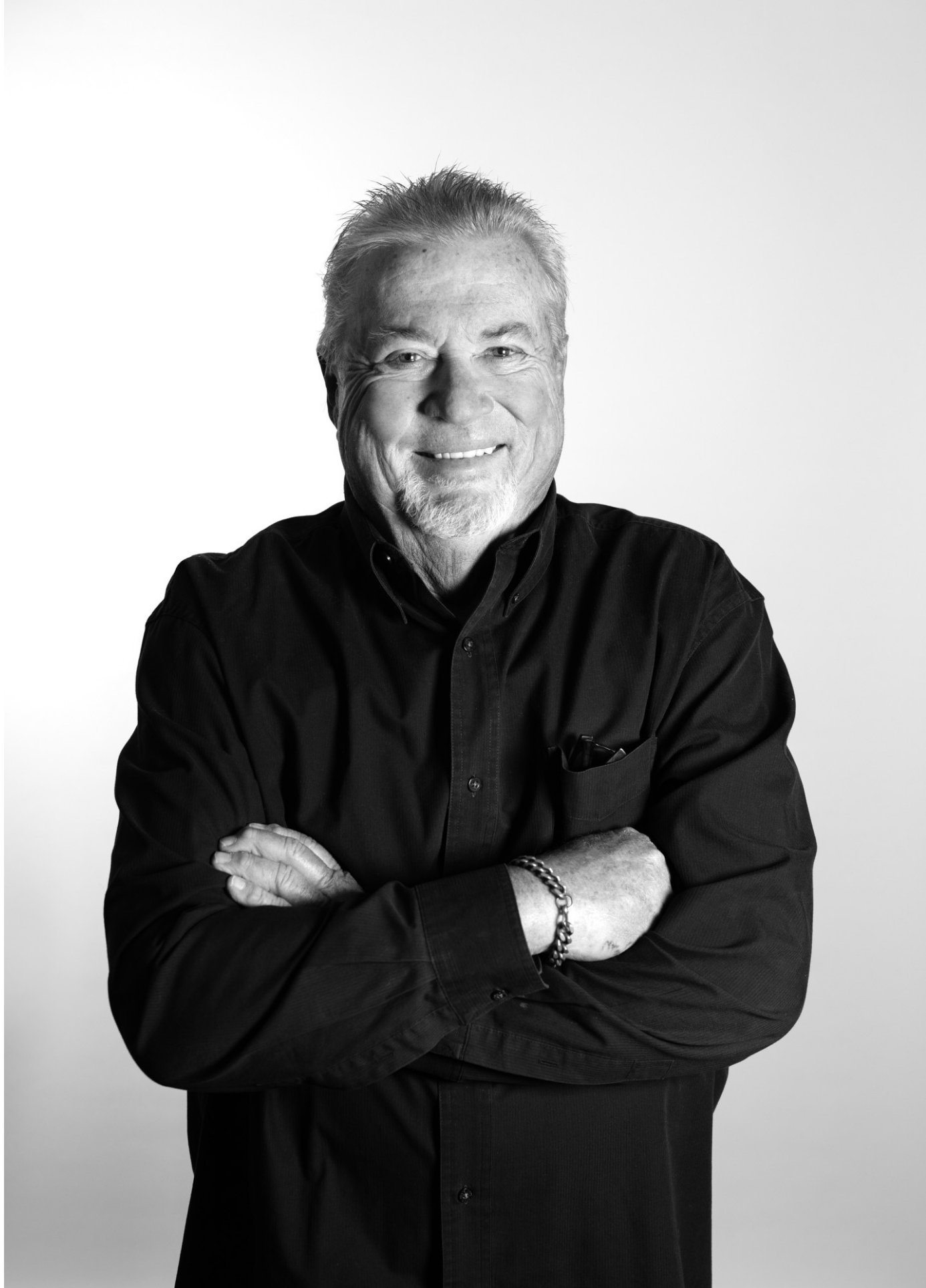 Black and white portrait of an elderly man with a beard and mustache, smiling, wearing a dark button-up shirt with sleeves rolled up, arms crossed, against a plain background.