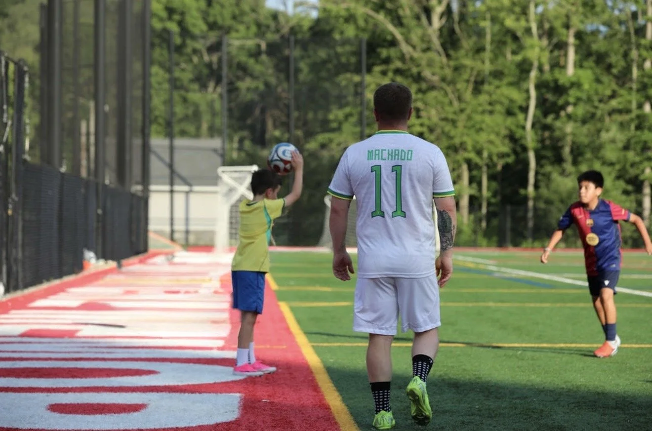 A soccer player in a white jersey with the name Machado and number 11 on the back playing with children on a soccer field, with one child preparing to throw a soccer ball.