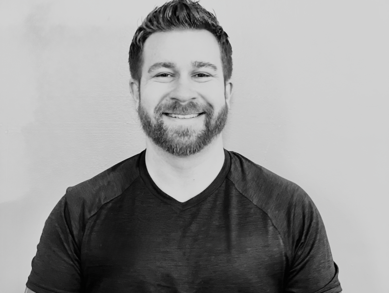 Black-and-white photo of a smiling man with a beard wearing a athletic t-shirt, standing against a plain wall.