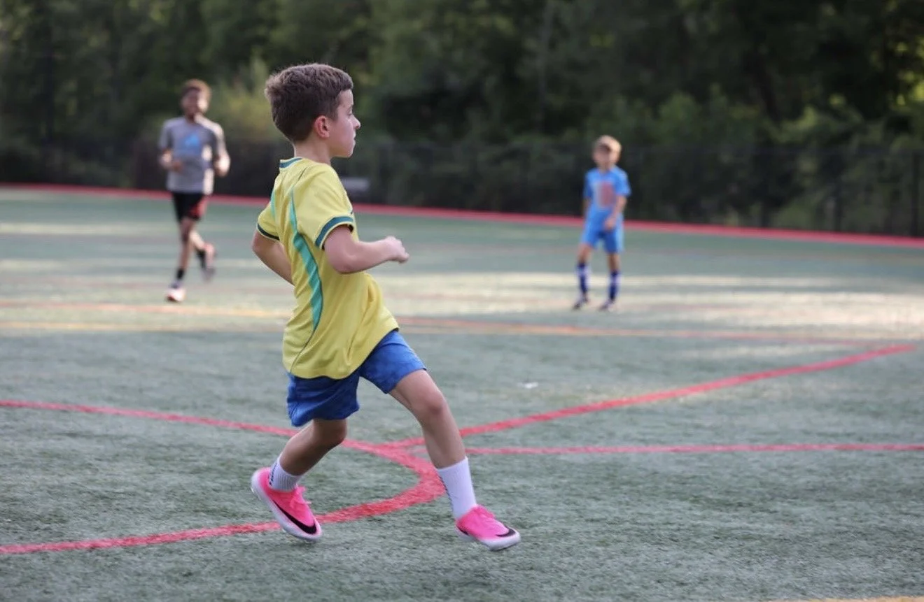 Children playing soccer on an outdoor field with trees in the background.