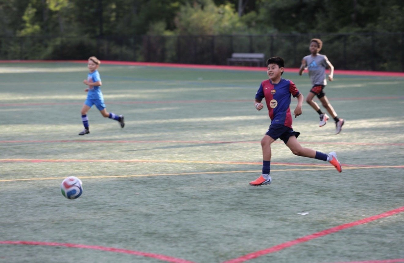Three children playing soccer on an outdoor field with trees in the background.