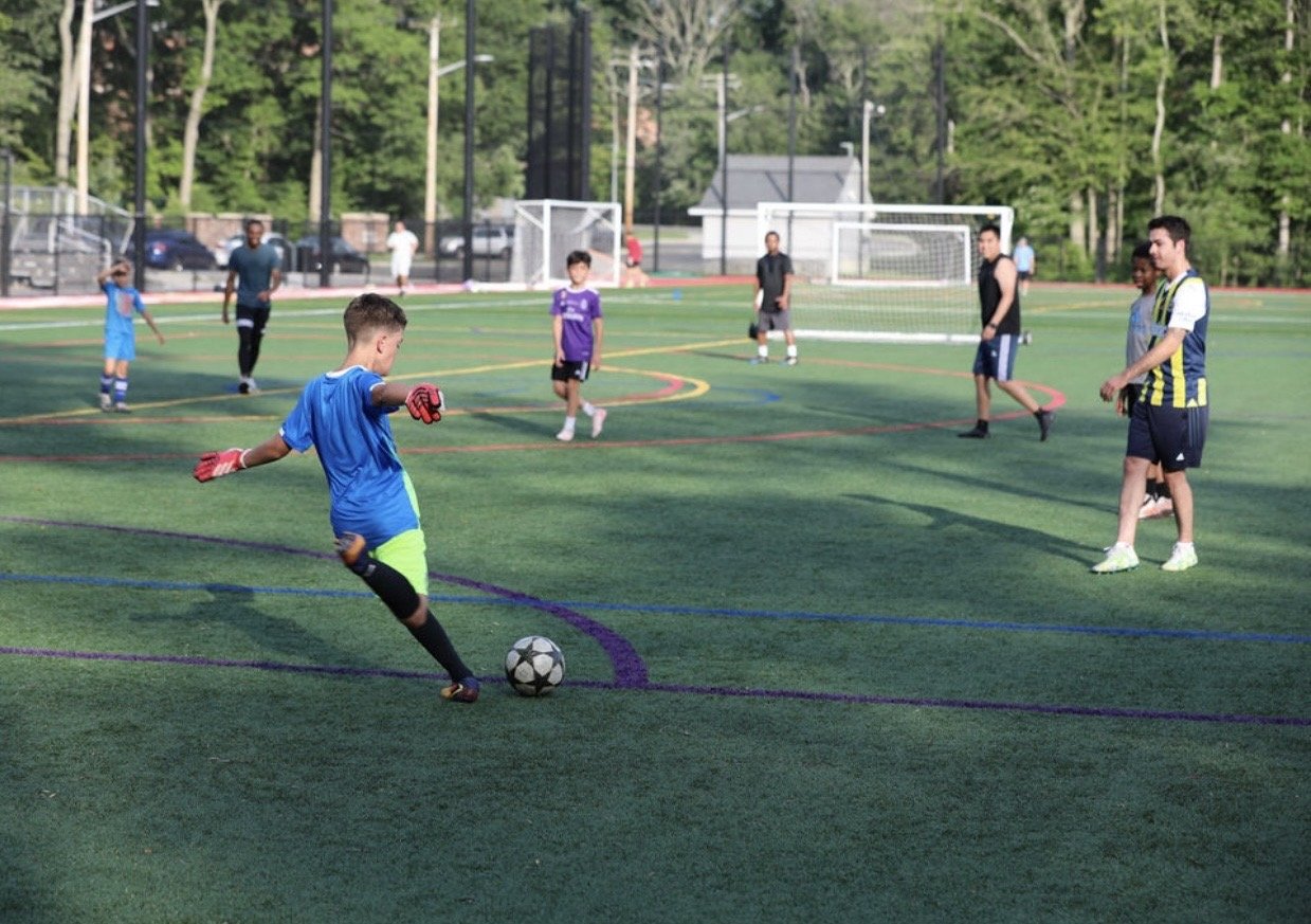 Young boys playing soccer on a green field during daylight, with one boy about to kick the ball while others watch or stand nearby.