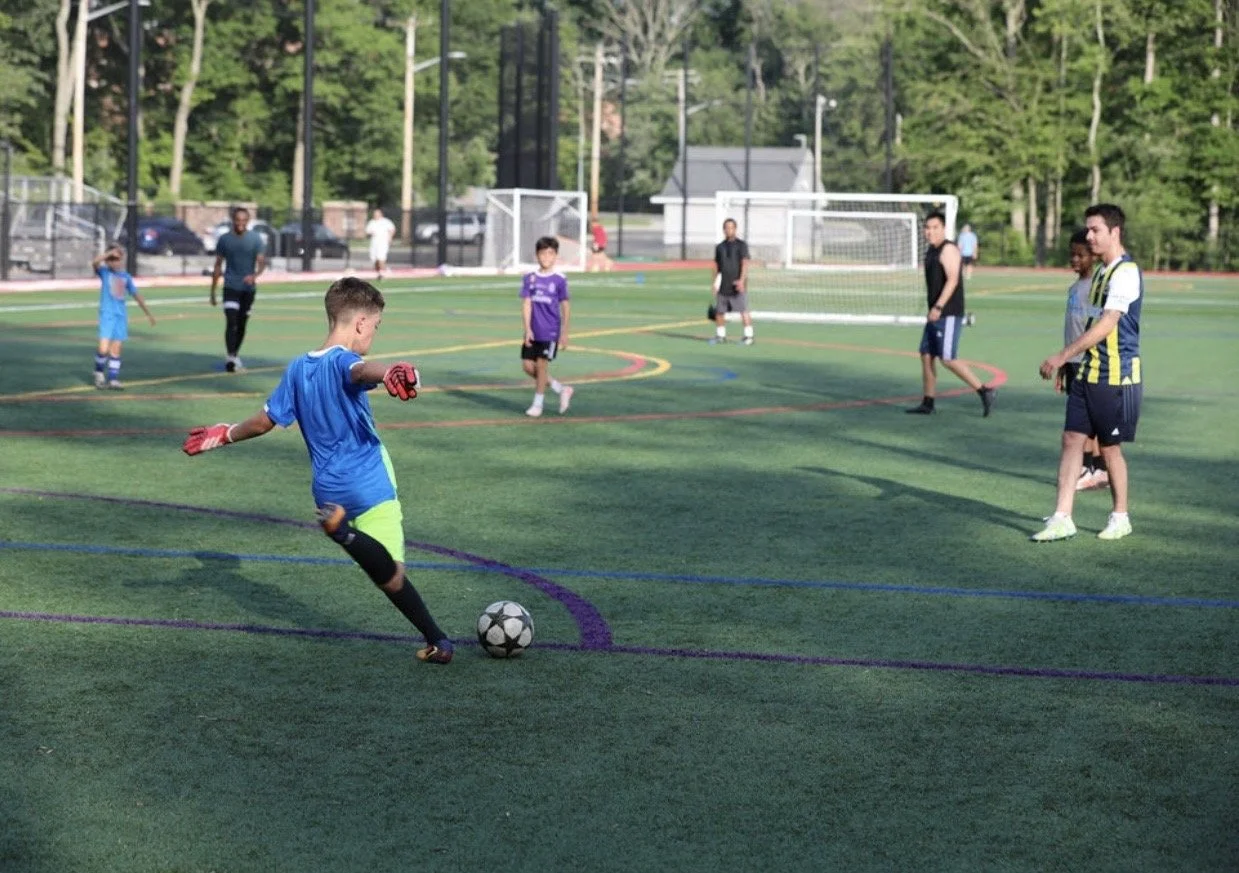 A group of kids and teenagers playing soccer on a green field with goal posts and trees in the background.