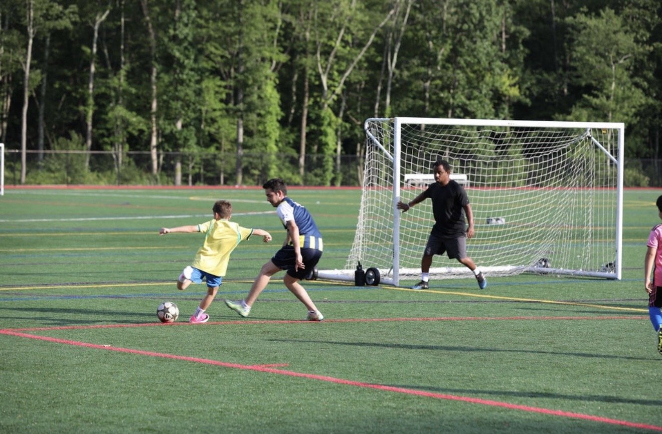 Two children and a teenager playing soccer on a field, with a goalkeeper in black preparing to block a shot, and trees in the background.