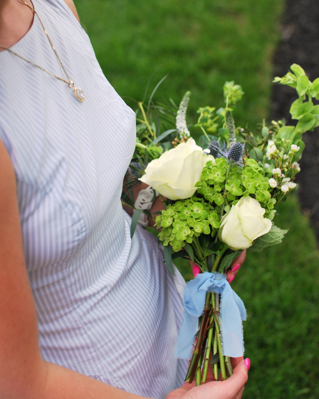 Person wearing a white dress holding a bouquet of white roses, green hydrangeas, and other flowers tied with a blue ribbon, standing on grass.