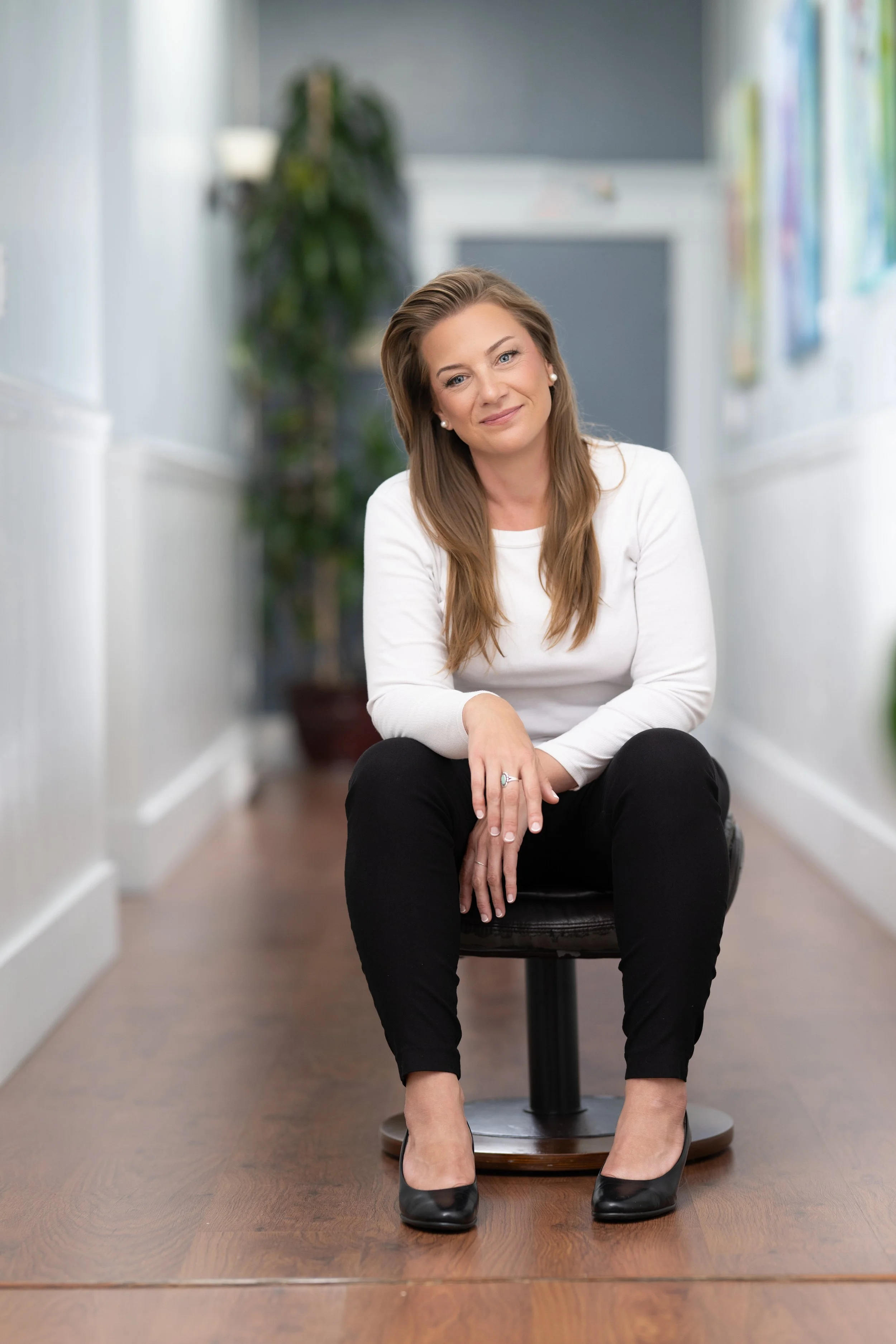 A woman with long brown hair sitting on a black stool in a hallway, wearing a white long-sleeve shirt, black pants, and black high heels, smiling at the camera with a blurred background of a hallway with artwork on the walls and a large green plant.