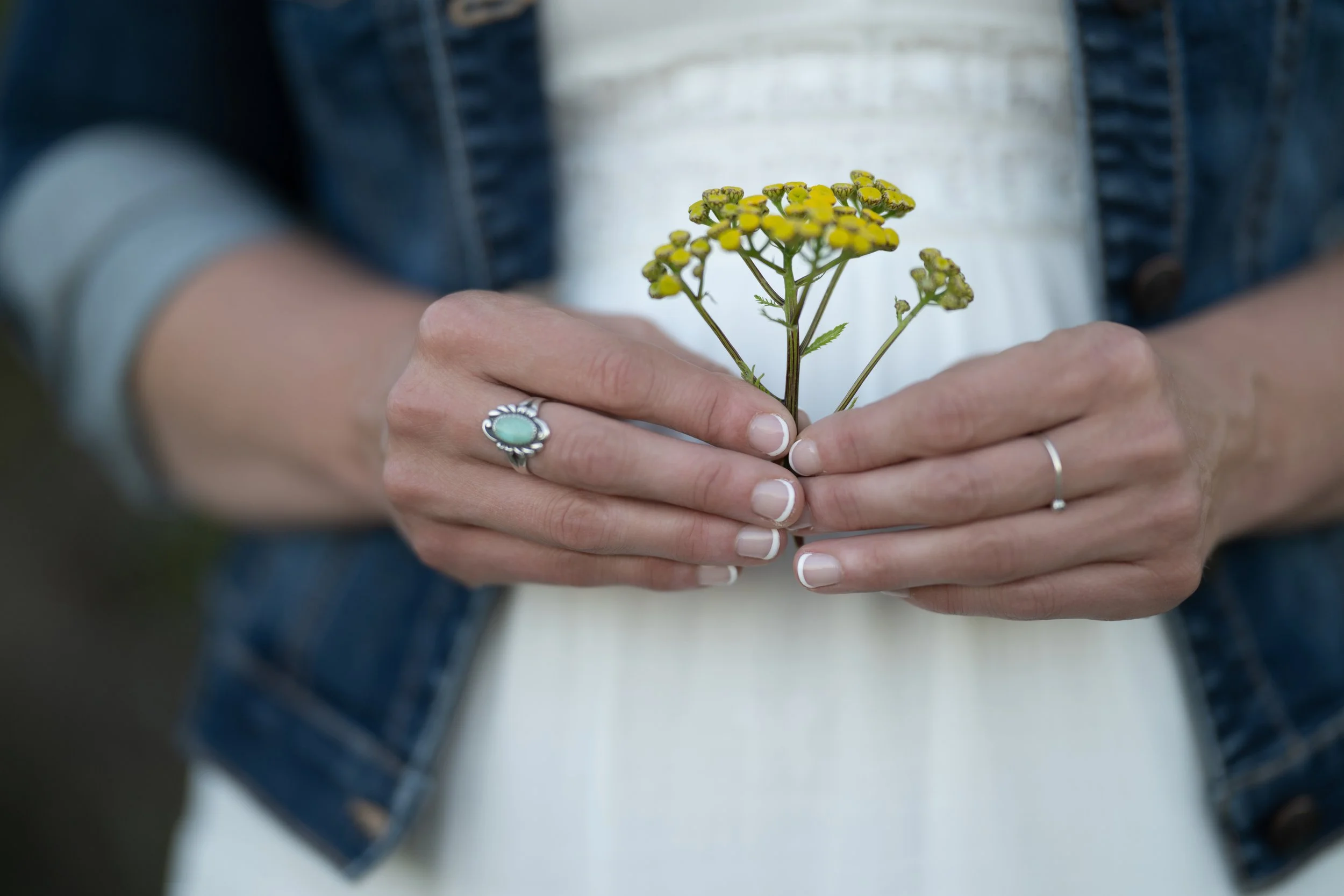 Renate Horst holding a small yellow flower with green stems and leaves, wearing rings on fingers, dressed in casual clothing including a denim jacket.