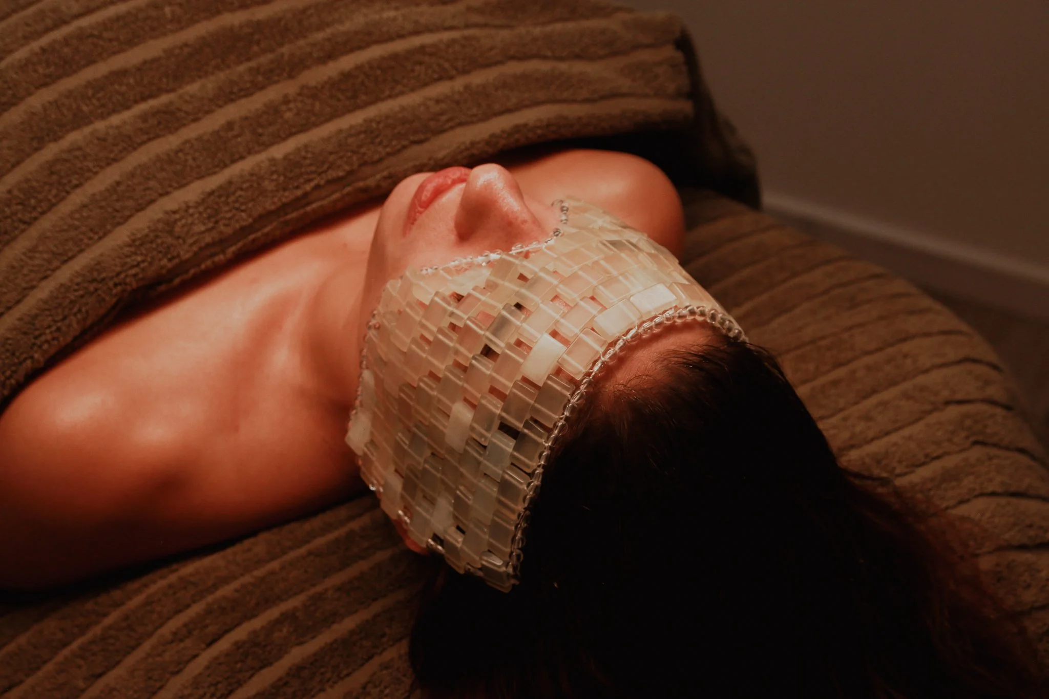 Person lying on their side on a brown textured couch, wearing a beige mosaic-patterned face mask and a brown striped long-sleeve shirt.