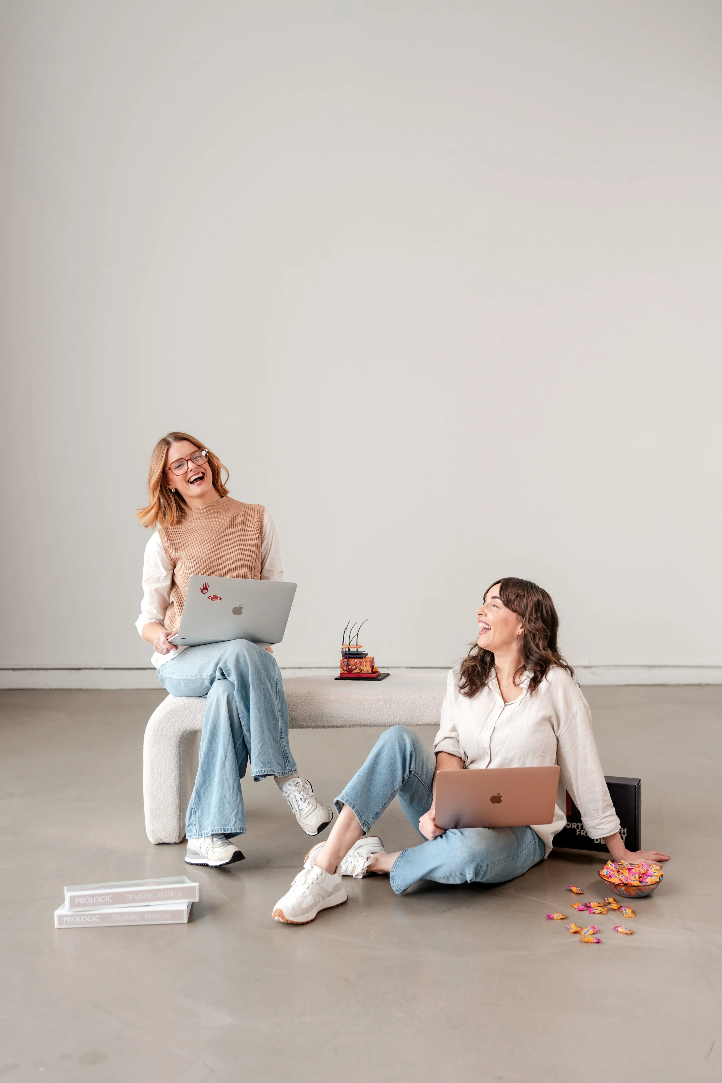 Two women with laptops sitting and standing on a bench against a plain wall, laughing and chatting, with colorful candies spilled on the floor nearby.