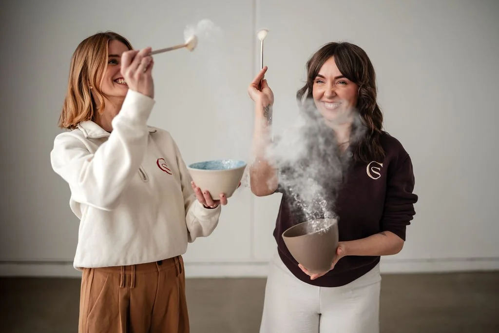 Two women are happily playing with dry ice, creating vapor smoke. One woman is holding a bowl, and the other is holding a mug, both with spoons. They are smiling and appear to be enjoying a fun activity together.