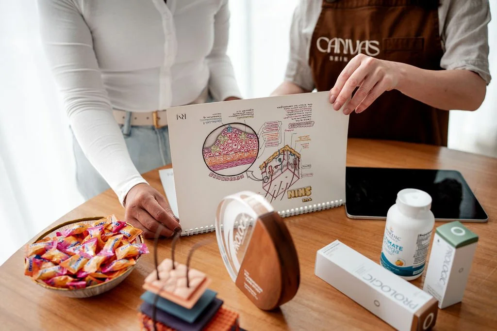 Two women sitting at a wooden table discussing scientific diagrams and samples, with medicine bottles, a tablet, and colorful fabric swatches on the table.