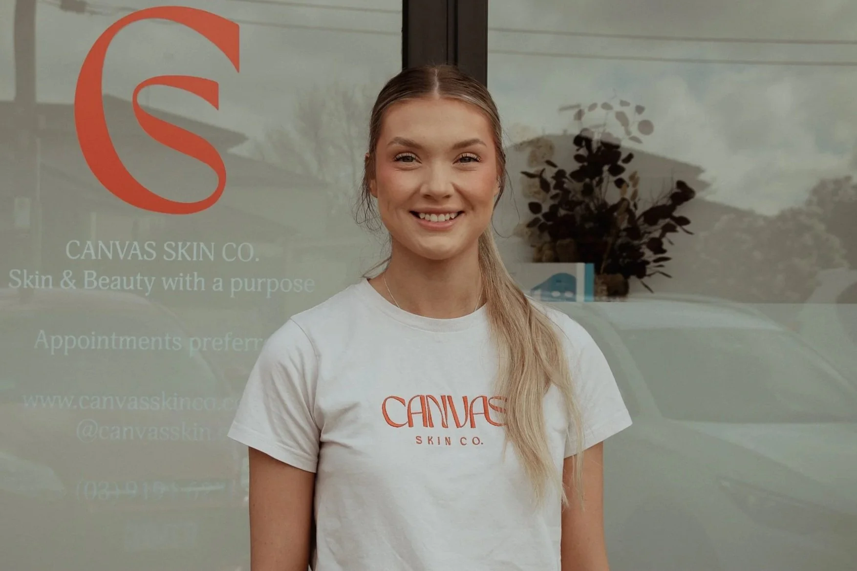A woman standing in front of a glass door with the logo and name of Canvas Skin Co. on it, smiling at the camera. She is wearing a white t-shirt with the company's logo.