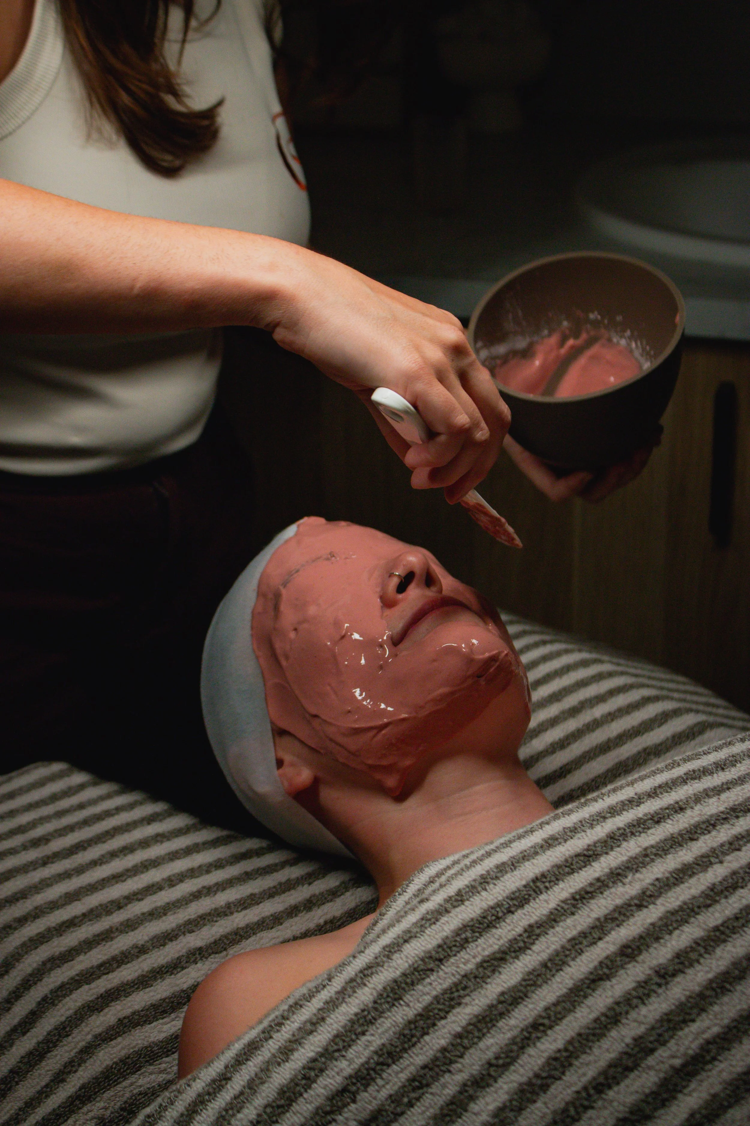 A person lying down with a facial mask, whose face is partially visible, while another person applies a facial mask to their face.