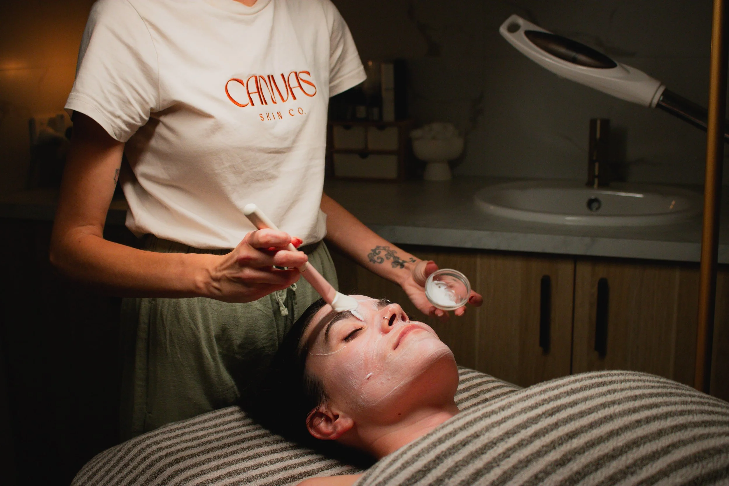 A woman receives a facial treatment while lying on a spa bed, with a skincare professional applying cream to her face in a dimly lit room.