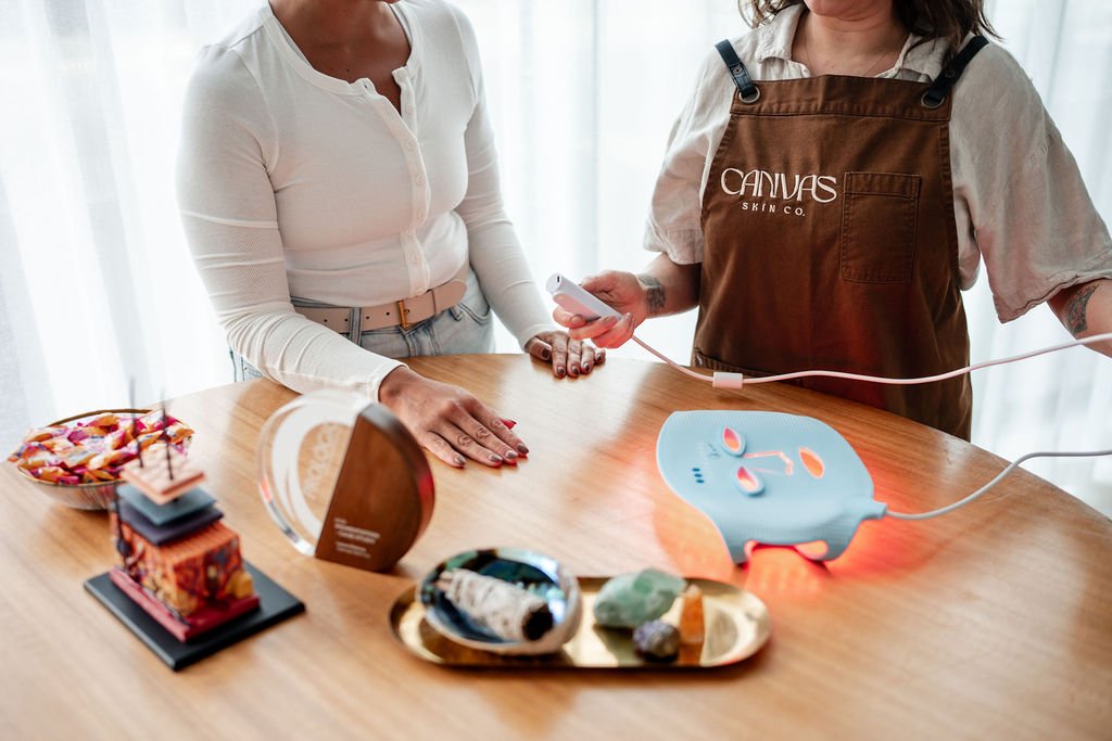 A woman receiving a facial treatment from an aesthetician using a red LED light therapy device connected to a control unit on a wooden table.