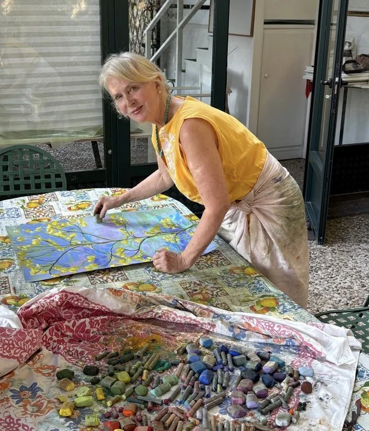A woman engaging in pastel art, painting a scene with tree branches and a sky, surrounded by colorful pastels on a table, in a room with large windows and a patterned tablecloth.