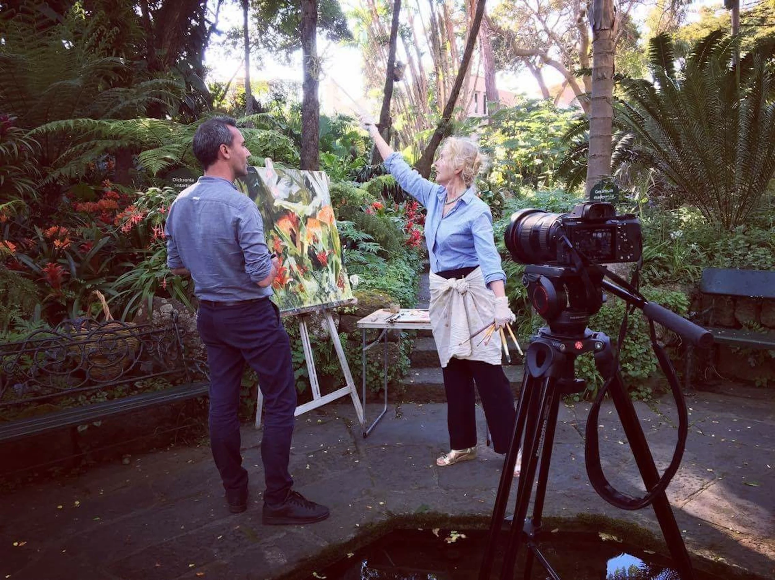 A woman shows a painting to a man in a lush, green garden with dense foliage and trees, with a camera set up in the foreground.