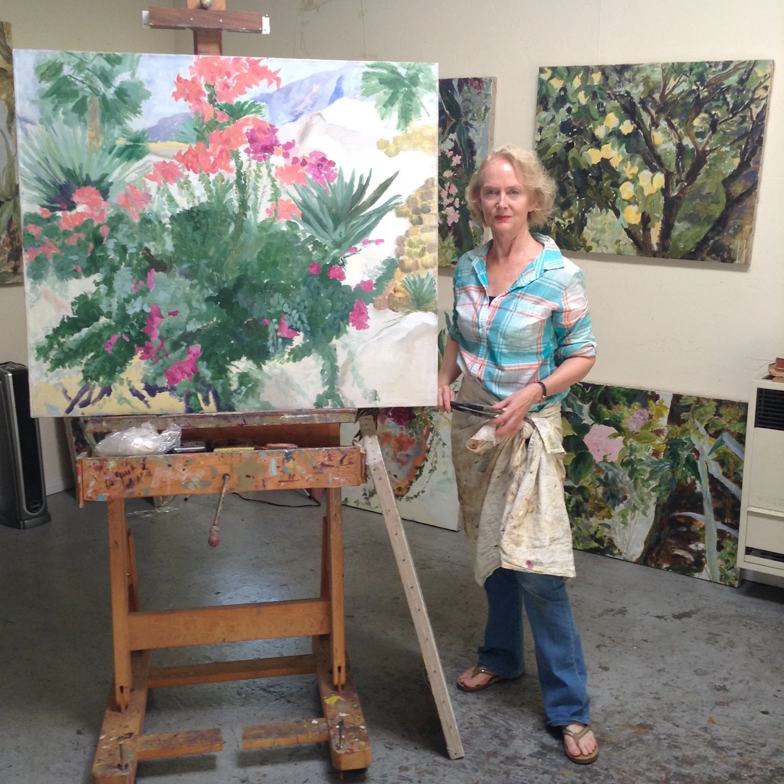 An artist standing in art studio next to large, unfinished floral landscape painting on canvas, with trees and colorful flowers, wearing a plaid shirt and sandals.