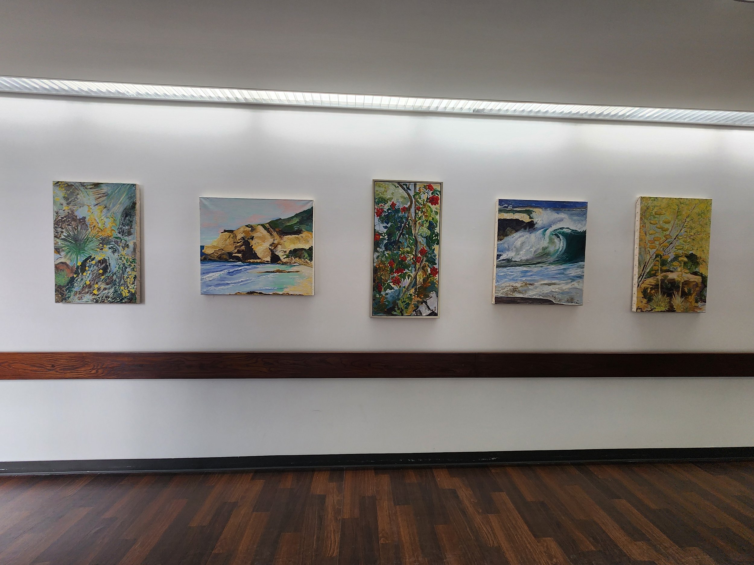 Five landscape paintings hanging on a white wall in an art gallery, with a dark wooden floor below.