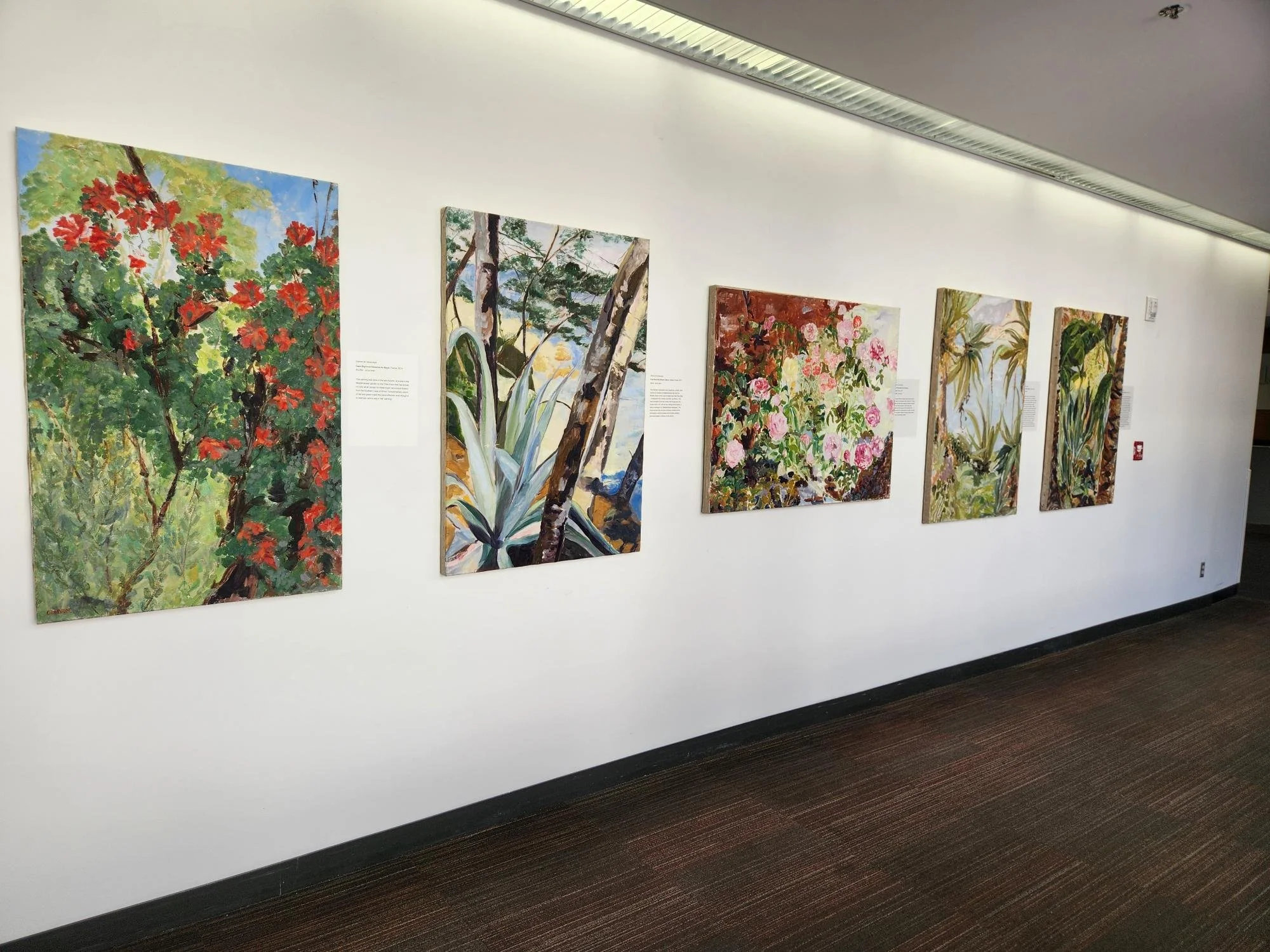 A row of colorful landscape paintings displayed on a white gallery wall. The paintings depict nature scenes with trees, flowers, and greenery, in a variety of styles and compositions.