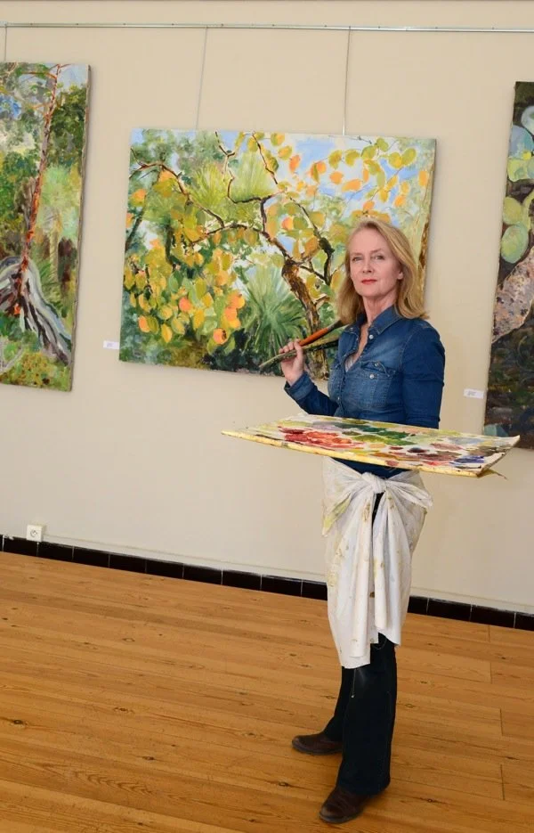A woman holding a painter's palette and brush stands in an art gallery with colorful landscape paintings on the wall.