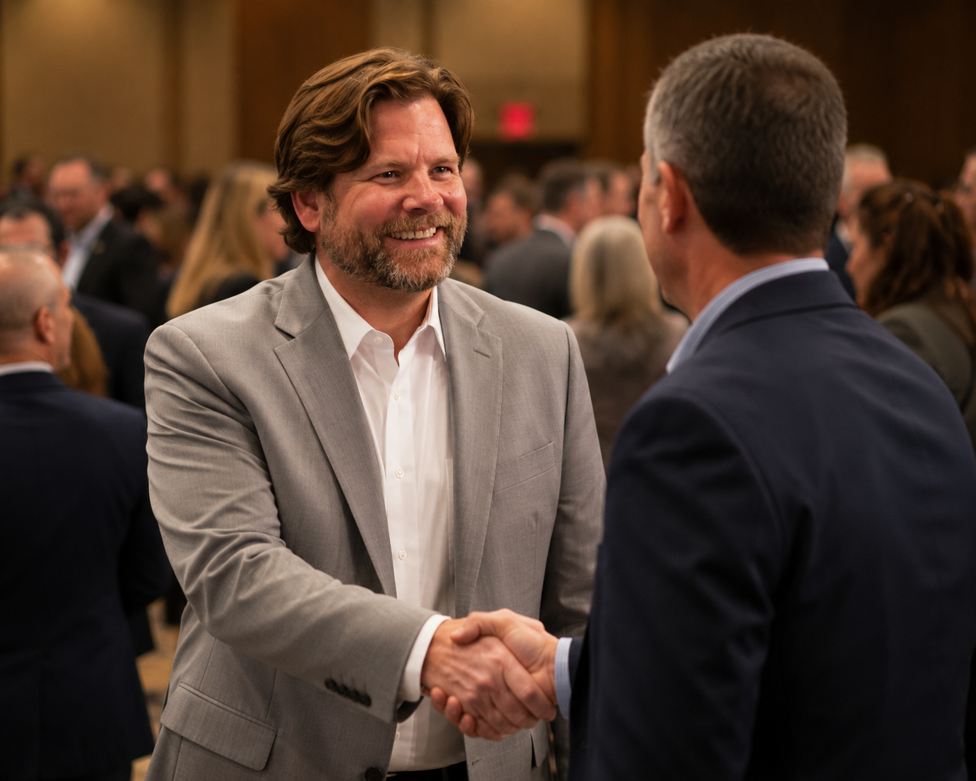 Two men in suits shaking hands at a formal gathering, with a crowd of people in the background.