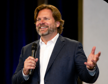 Man with brown hair and beard smiling, holding a microphone, wearing a dark suit and white shirt, speaking on stage.