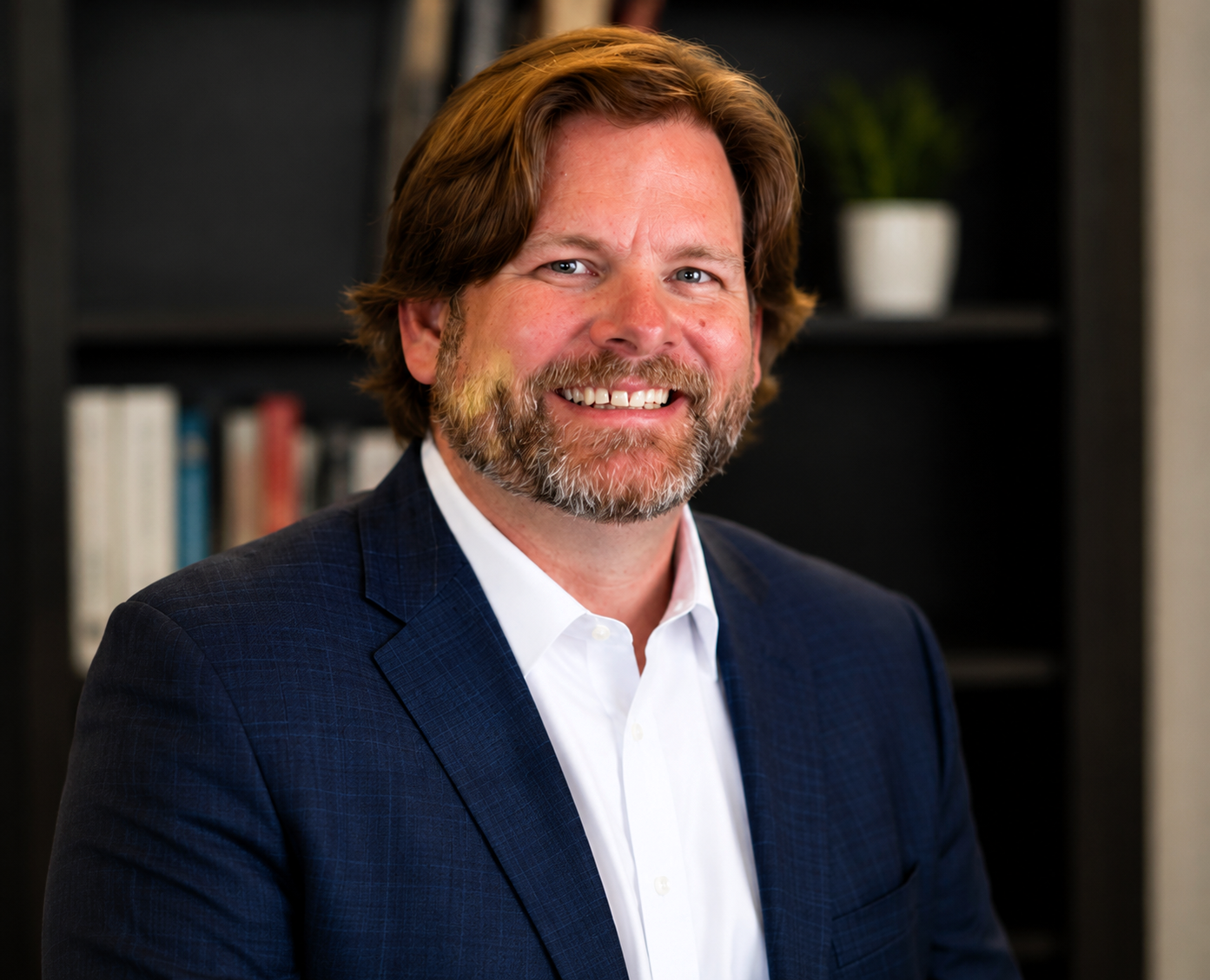 A smiling man with a beard and shoulder-length hair, wearing a navy blazer and white shirt, standing in front of a bookshelf with books and a potted plant.