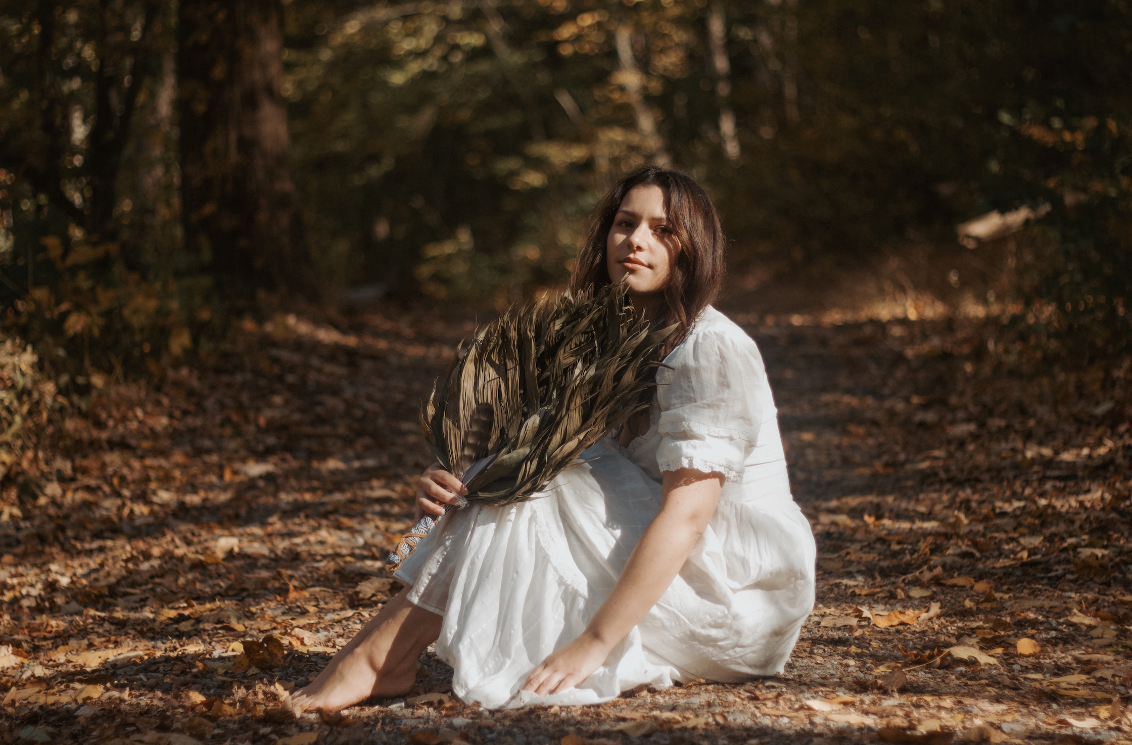 A young woman with wavy brown hair wearing a white dress, sitting on fallen autumn leaves in a forest, holding a bundle of dried leaves, with trees and sunlight in the background.