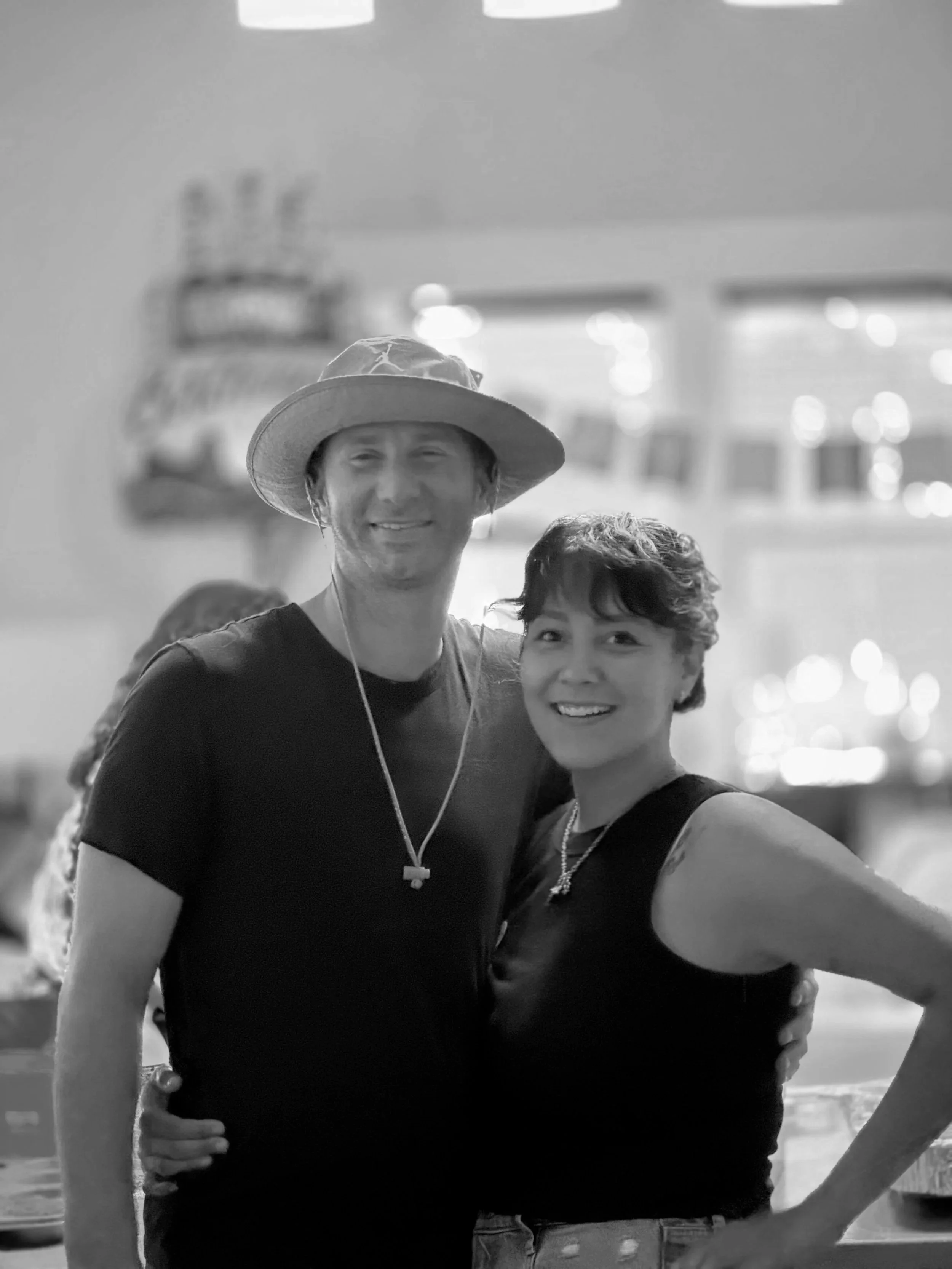 Two smiling women, one wearing a wide-brimmed hat, standing close together indoors with blurred background that includes windows and lights.