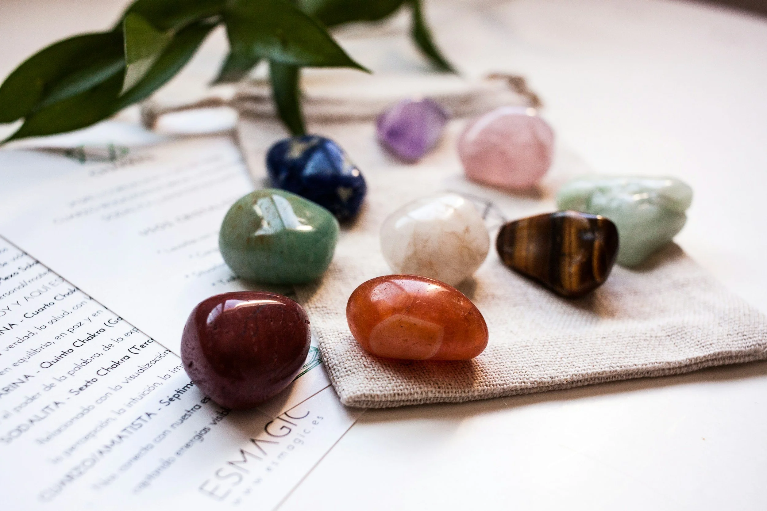Colorful polished stones arranged on a beige fabric cloth, with some green leaves and partially visible printed papers underneath.