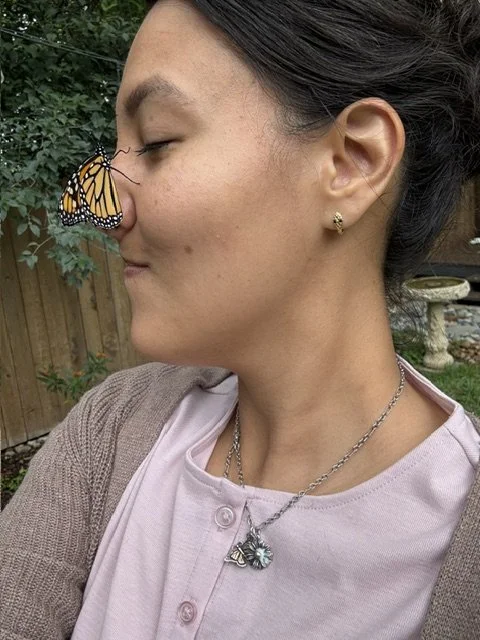 A woman with dark hair and small earrings, wearing a light pink shirt and a necklace, has a butterfly resting on her nose.