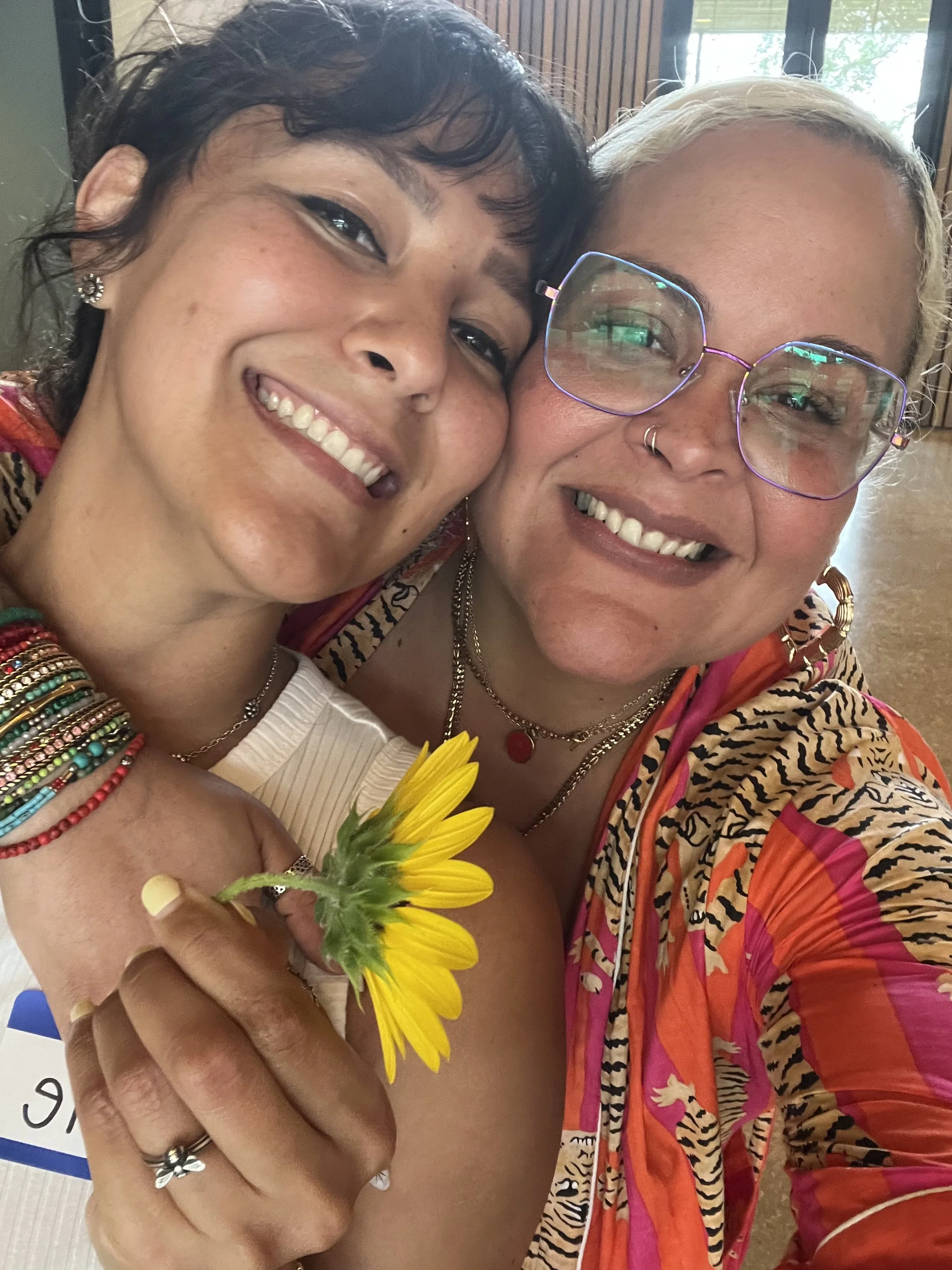 Two women smiling and hugging, one woman holding a sunflower. The woman on the right has light blonde hair, large glasses, and a nose ring. The woman on the left has short curly dark hair and colorful beaded bracelets. They are indoors with windows in the background.