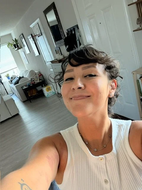 A woman taking a selfie in a bright living room. She has short, curly dark hair, is wearing a white sleeveless top and jewelry, and is smiling at the camera.