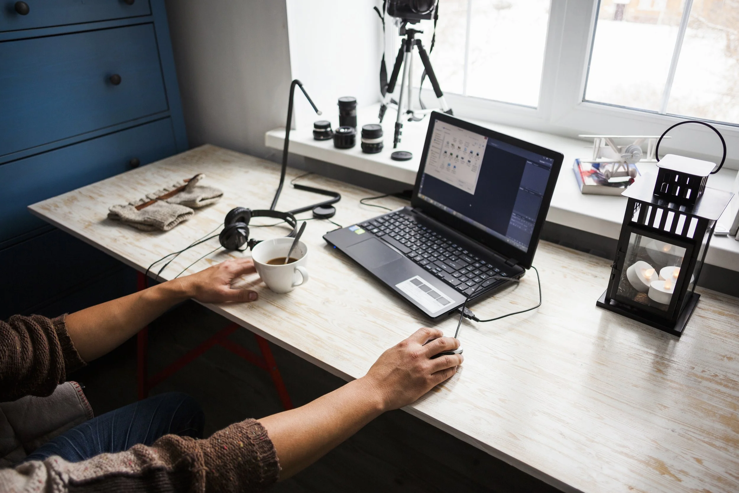 Inside a home office with a wooden desk near a window, a person wearing a brown sweater is using a computer mouse. On the desk are a laptop, headphones, a coffee mug, and various camera lenses. The window has a camera on a tripod and a small tray with headphones and small decorative candles.
