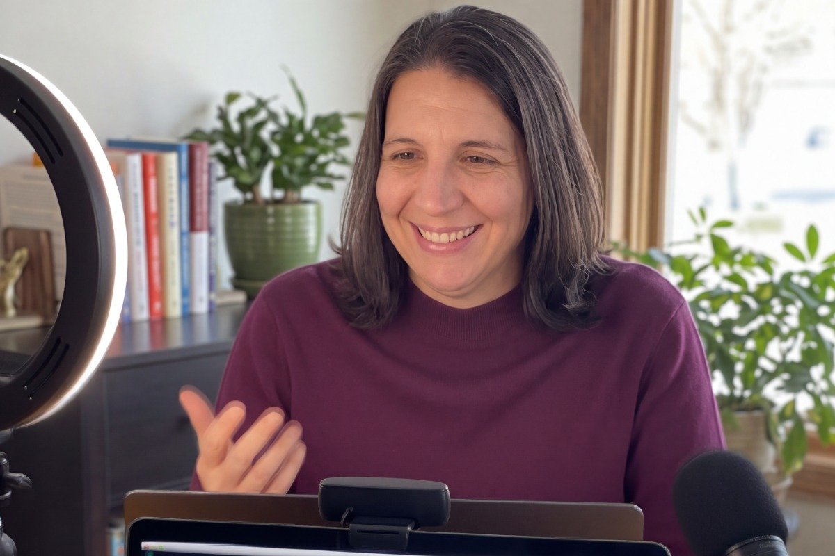 A woman with shoulder-length dark hair smiling during a video call or recording session, surrounded by a ring light, a laptop, and plants, with a bookshelf in the background.