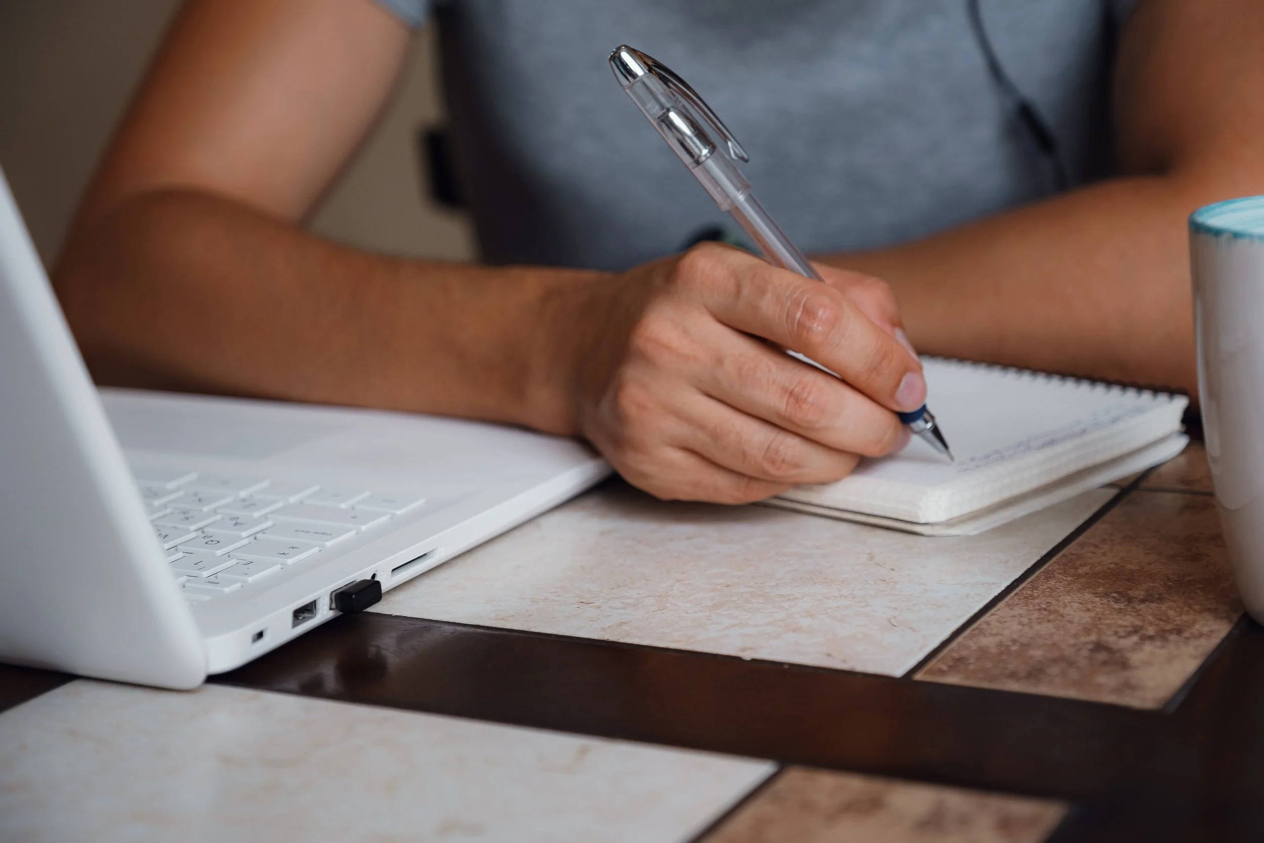 Person writing in a notebook with a pen at a desk, with a laptop nearby and a mug on the right.