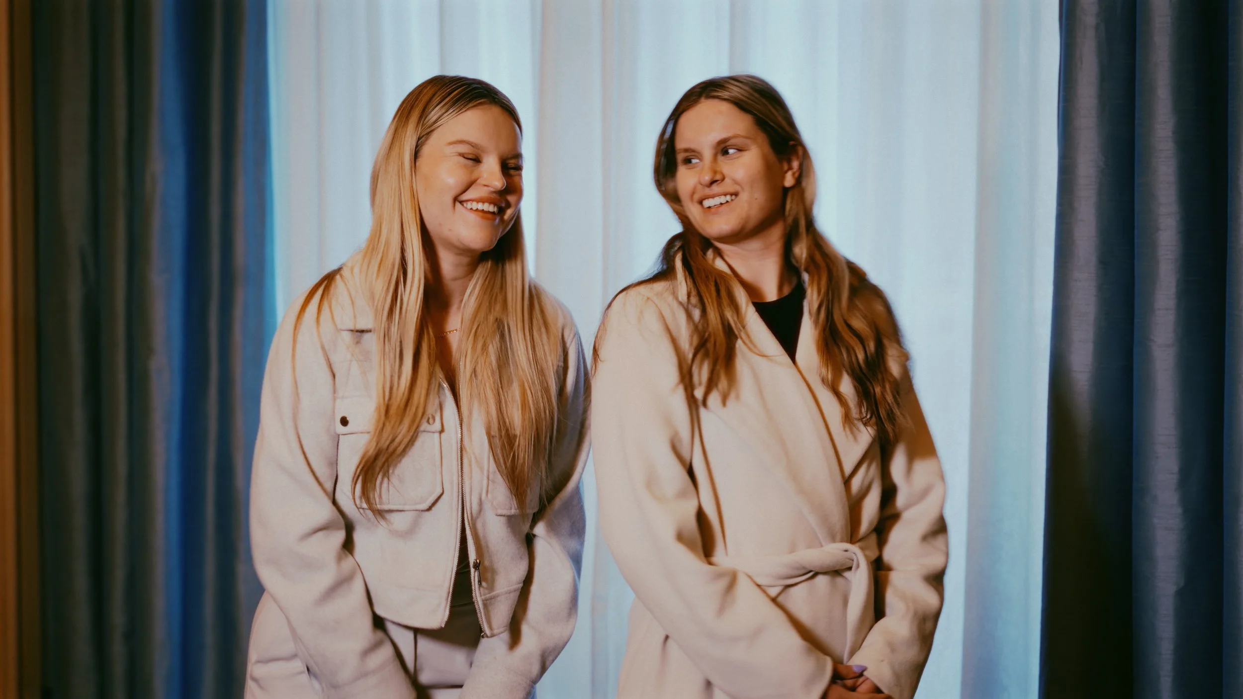 Two women standing near curtains, smiling and looking at each other.