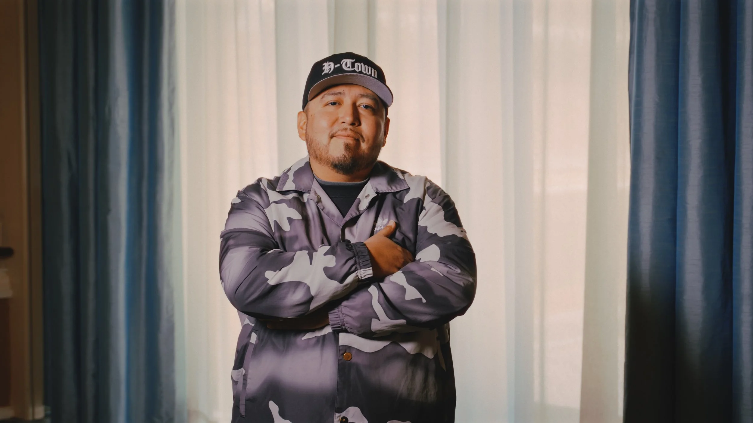 A man with a beard and mustache wearing a black cap with white lettering that reads 'H-Town', a camouflage jacket, and black shirt standing with arms crossed in front of a background of light-colored curtains.