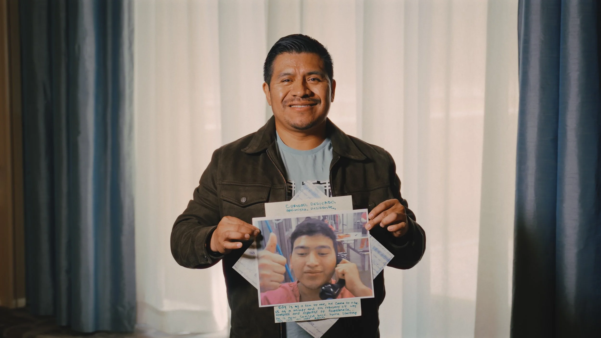 A smiling man with dark hair holding a photo of a young boy. The photo has handwritten notes at the top and bottom. Background features curtains.