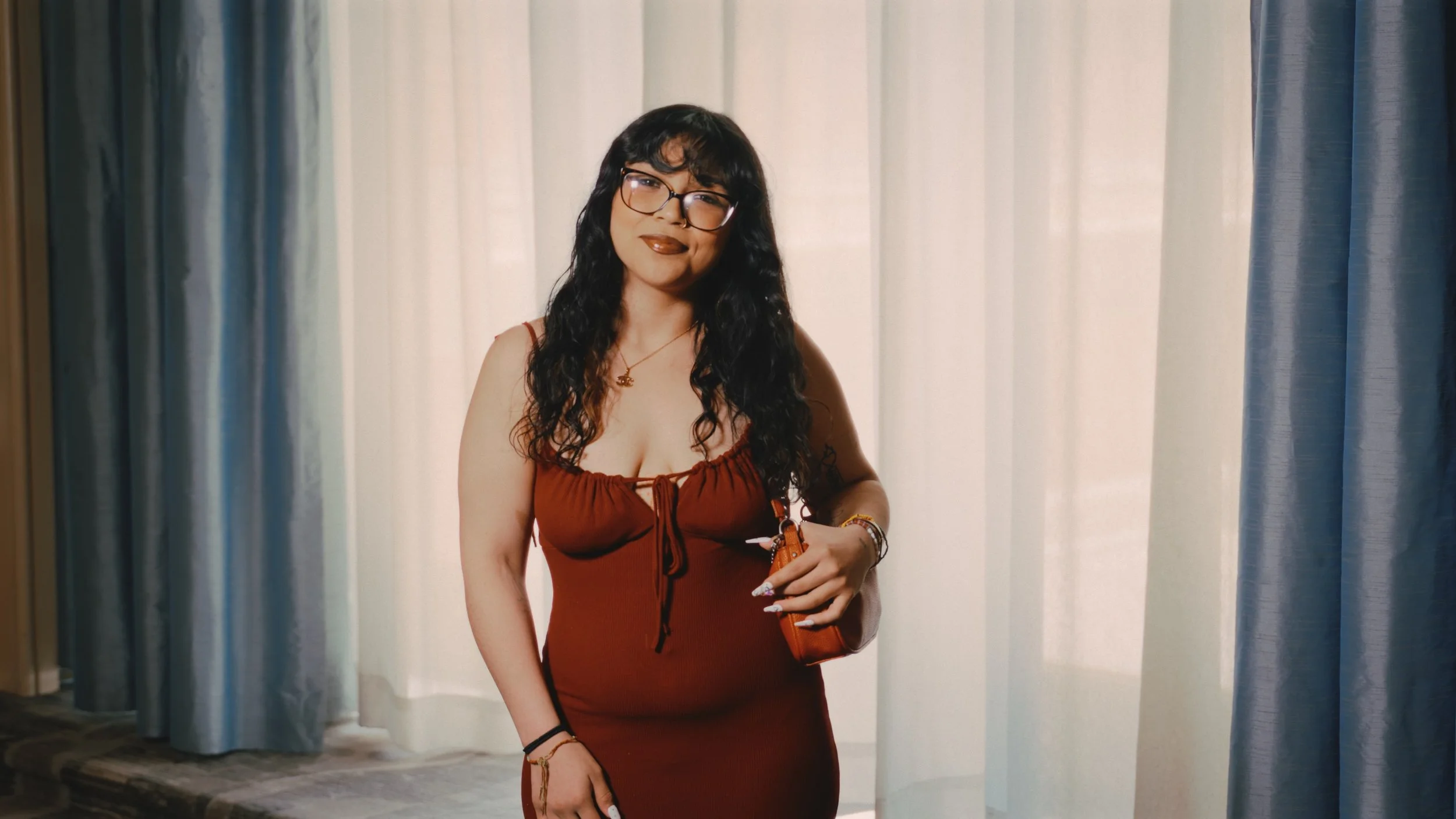 A woman with long curly black hair and glasses stands in front of light-colored curtains, wearing a red dress and accessorized with jewelry, smiling while holding a small orange bag.
