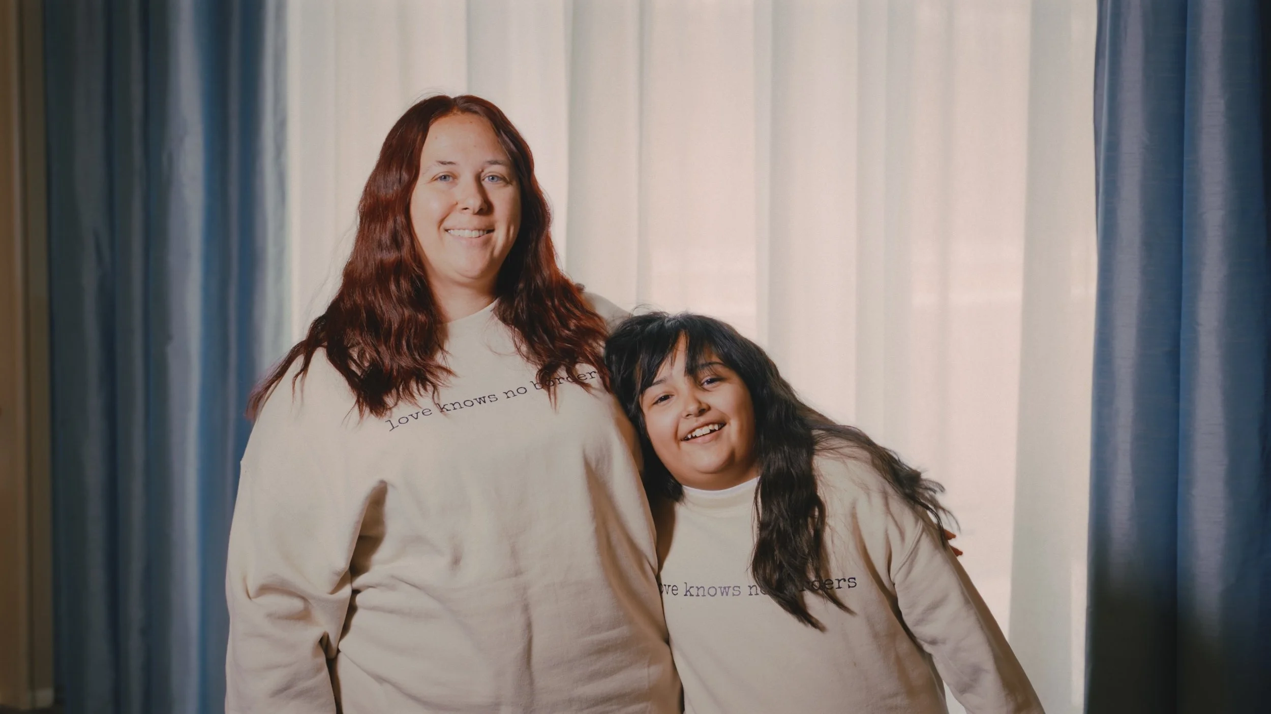Smiling woman with red hair and a young girl with long dark hair standing together in front of curtains, both wearing beige sweatshirts with the phrase "love knows no borders".