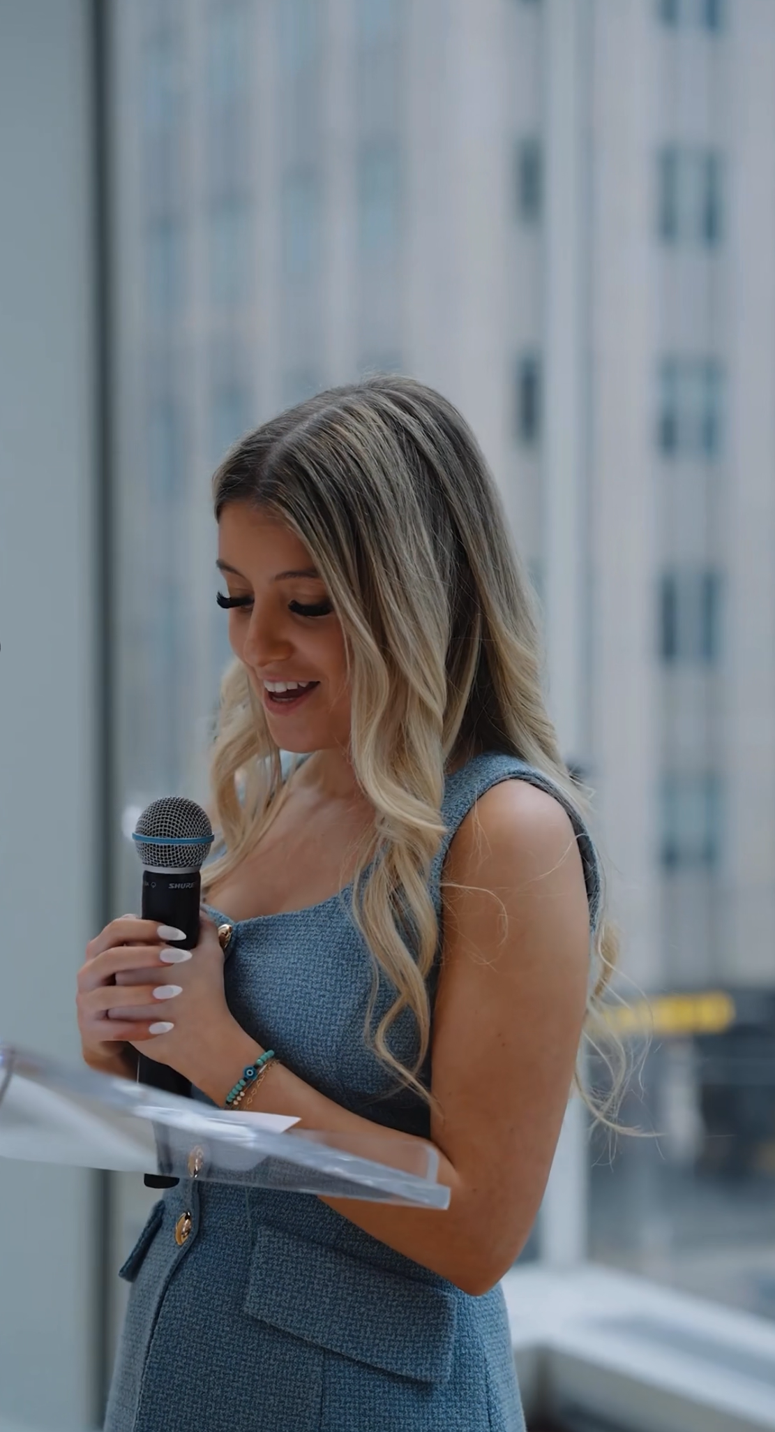 A woman with long blonde hair, wearing a blue dress, holding a microphone and speaking or singing indoors with a large window behind her.