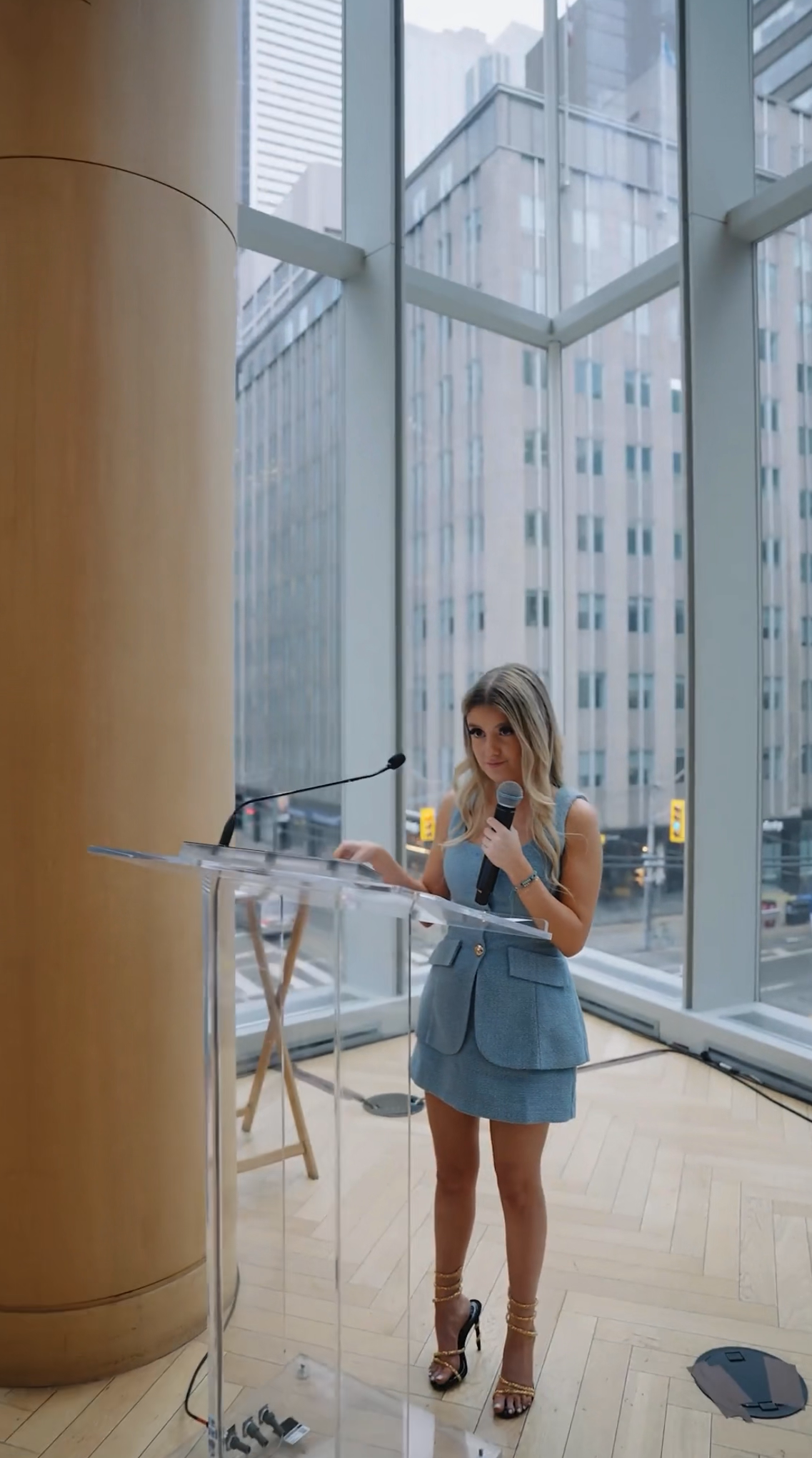 A woman in a blue dress and high heels standing at a clear podium, holding a microphone, in a modern room with large floor-to-ceiling windows showing tall buildings outside.