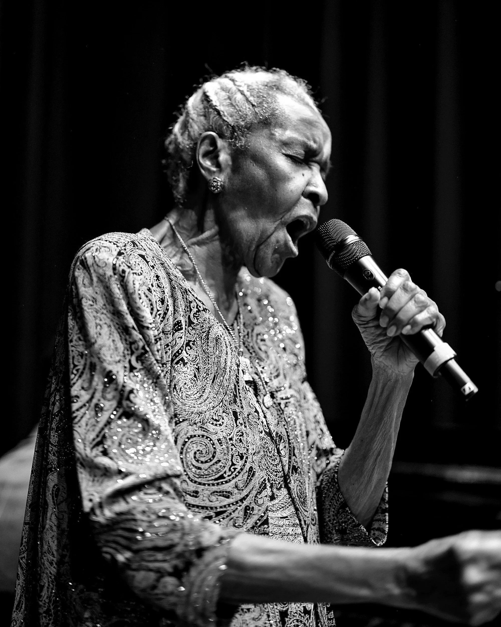 An elderly woman singing into a microphone on stage, wearing an ornate, patterned dress and jewelry, with a dark background.