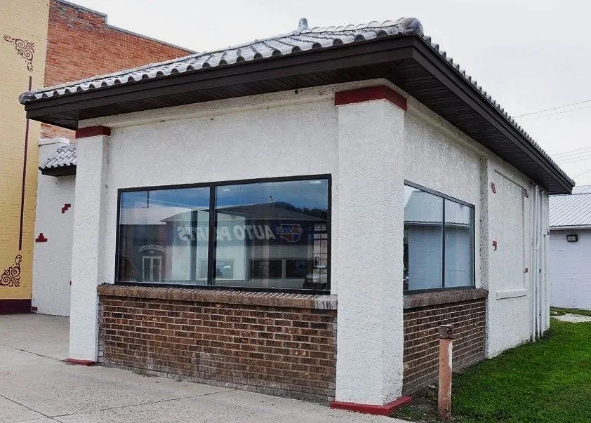 A small detail on a commercial. building. This pop-out, with a white stucco exterior and red brick lower siding, was formerly the gas station drive-through and has now been enclosed, featuring large windows and a dark metal overhanging roof.