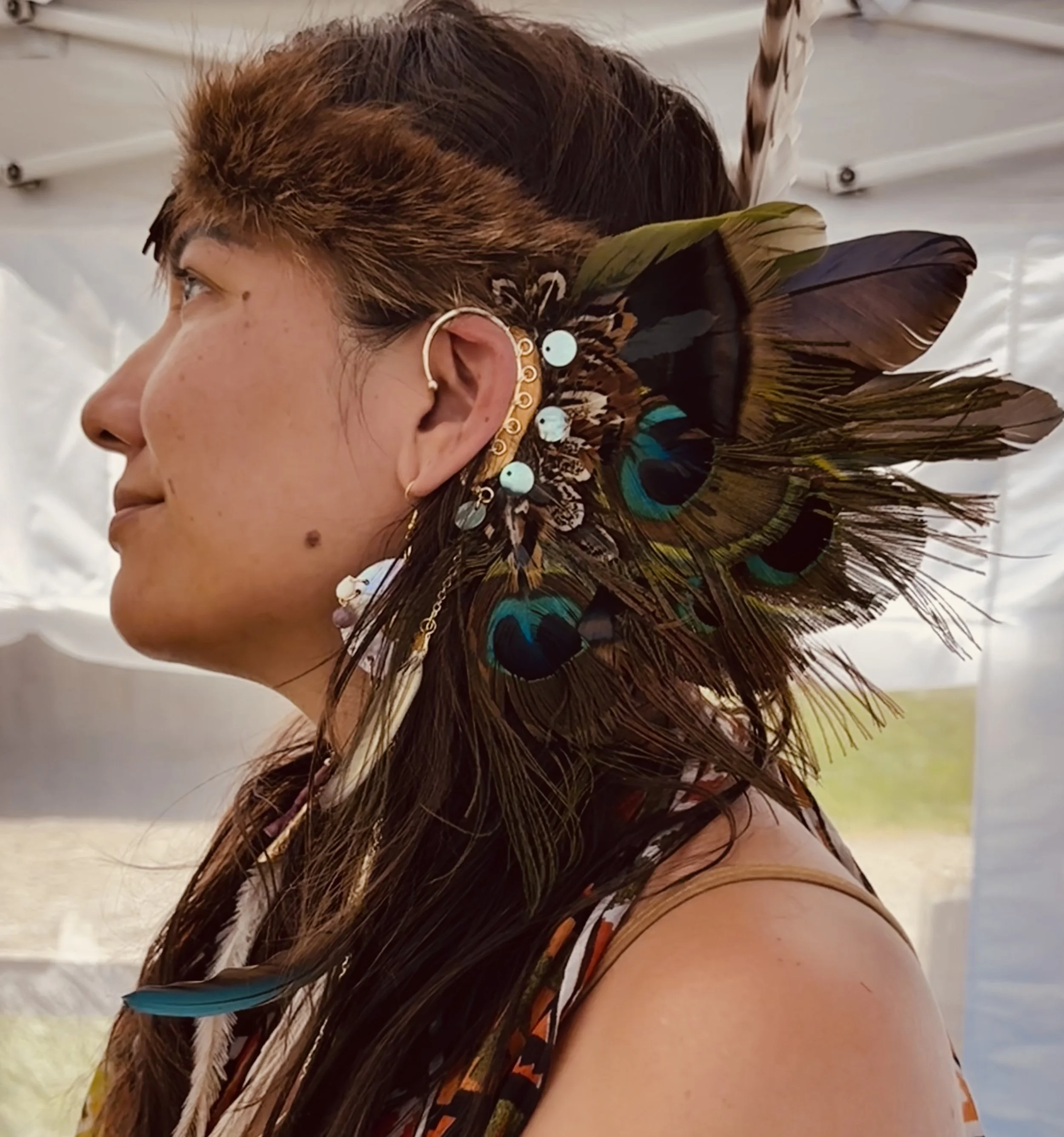 Close-up side profile of a woman with long brown hair decorated with peacock feathers, earrings, and a necklace, against a neutral background. This one of Cabane"s Native high-end jewelry artists.