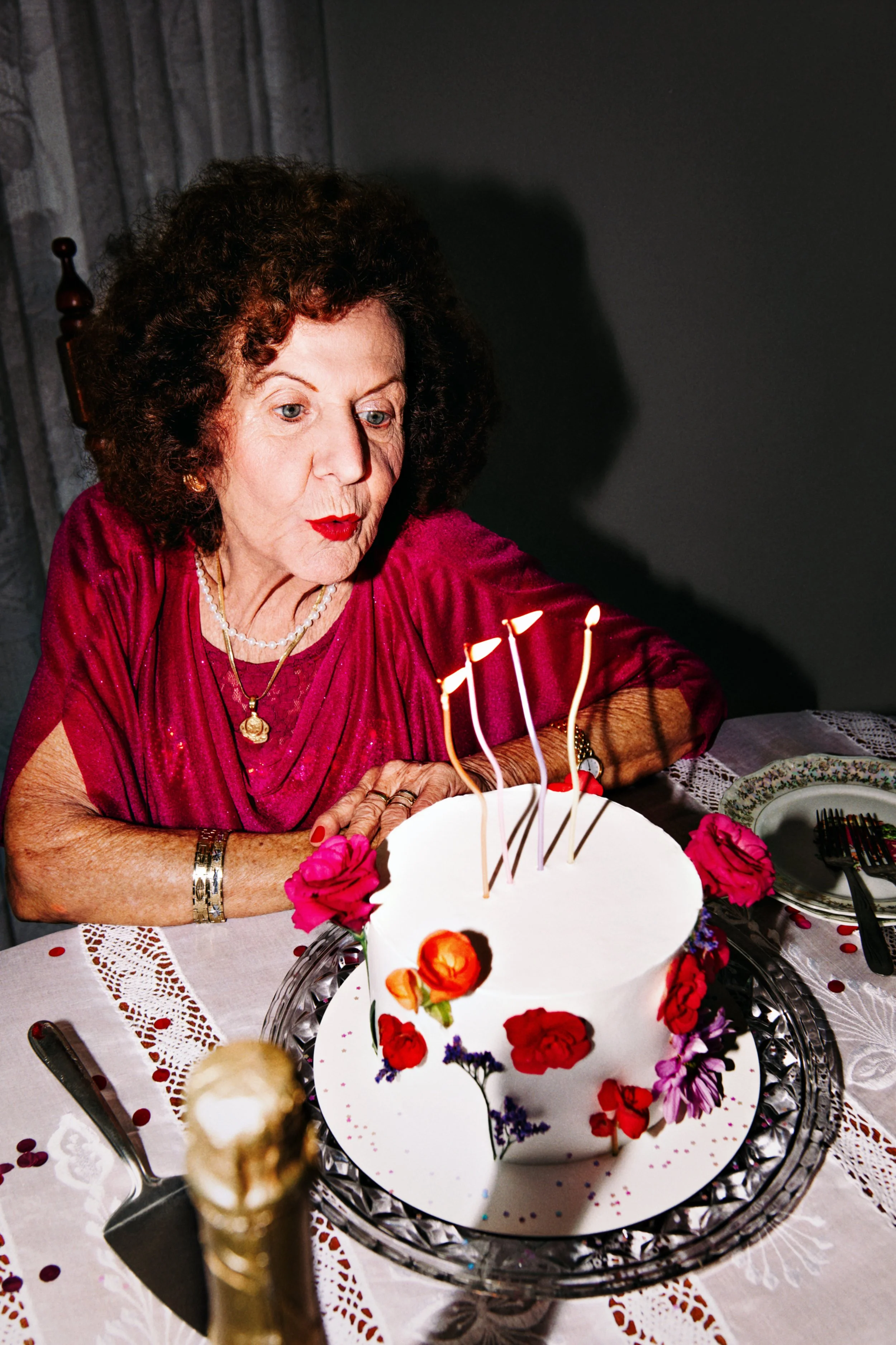 Fotografia com textura de filme 35mm e flash direto. Uma senhora de 80 anos, branca com olhos azuis, cabelo volumoso e cacheado em tom acobreado; usa um vestido magenta e joias douradas, inclina-se para soprar velas em bolo branco com flores reais.