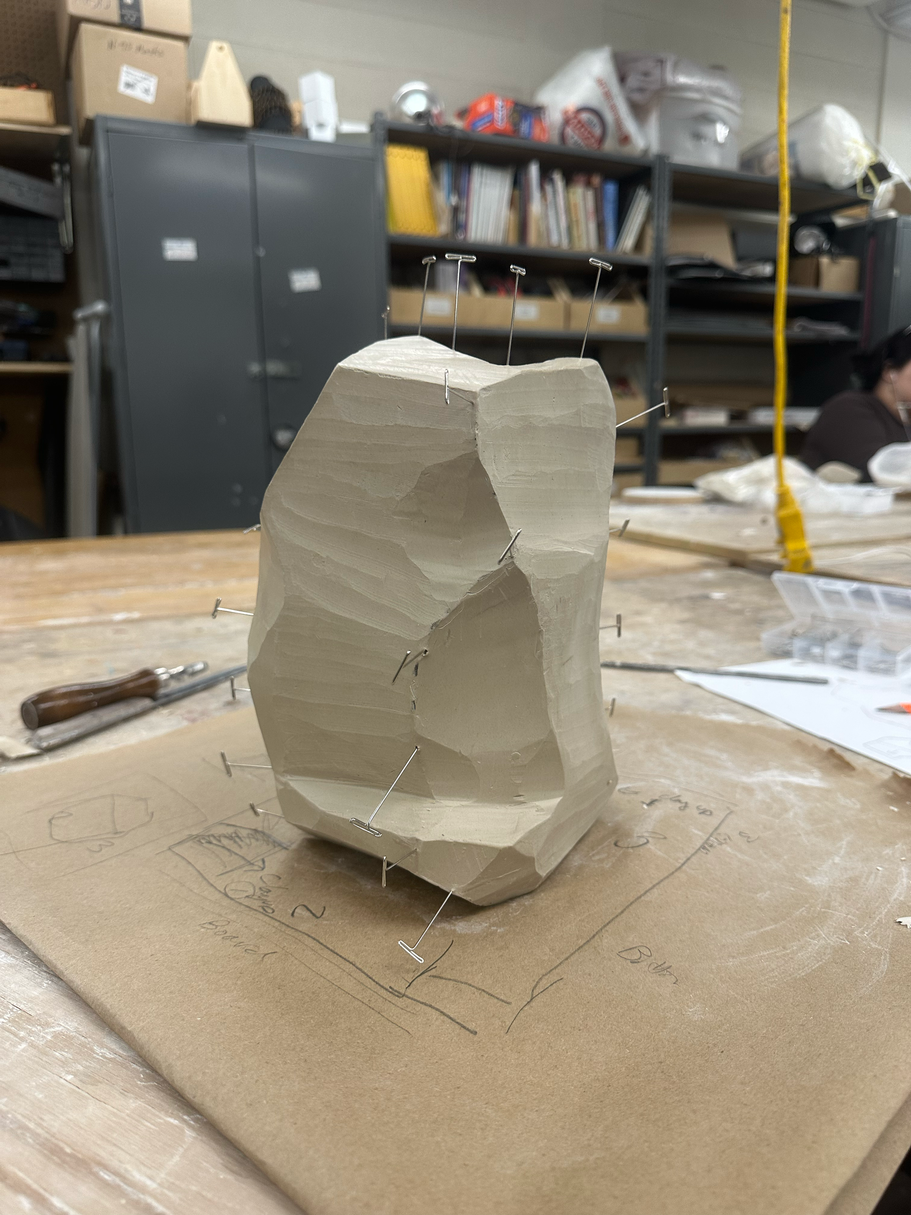 A carved piece of material secured with multiple pins on a worktable in a workshop or studio environment, with workspace and shelves in the background.