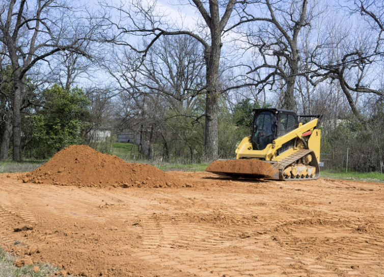 A small yellow bulldozer working on a dirt patch with a pile of dirt nearby, trees without leaves in the background.